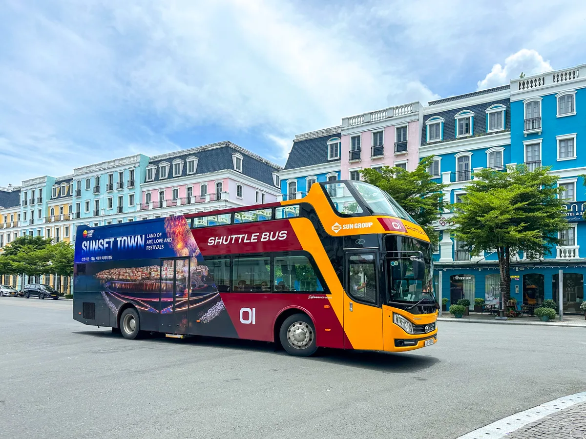 SunWorld double-decker shuttle bus parked in front of Sunset Town's colorful waterfront buildings